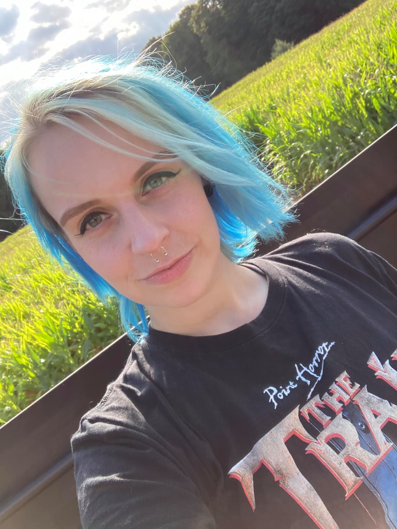 ADHD: A woman with blue shoulder length hair faces the camera wearing a black t-shirt with lettering on it. She is sitting in a corn field