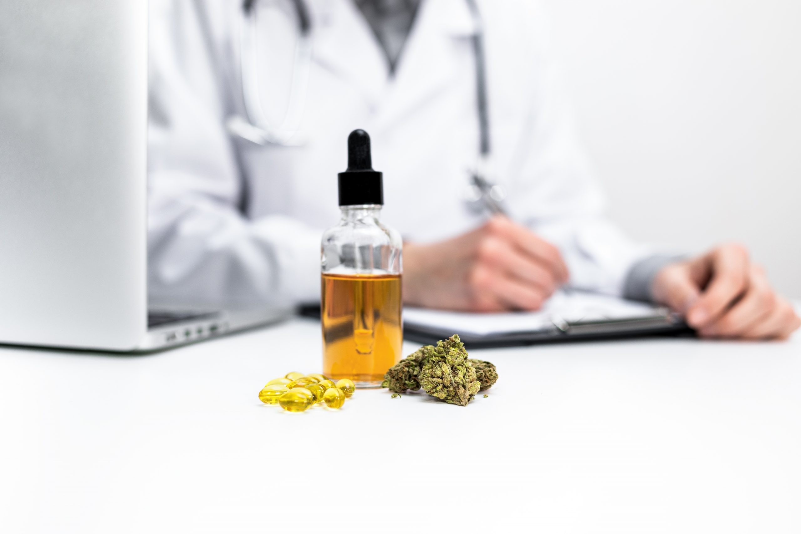 Medical Cannabis Prescriptions: A doctor in a white lab coat signed a prescription. In front of him, there is a yellow bottle with oil in it next to CBD capsules and a cannabis bud